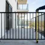 Garde-corps noir sur balcon d’immeuble moderne, mesureur jaune à côté, cadrage extérieur, barrière et façade blanche. Obligation mise aux normes garde corps évoquée.